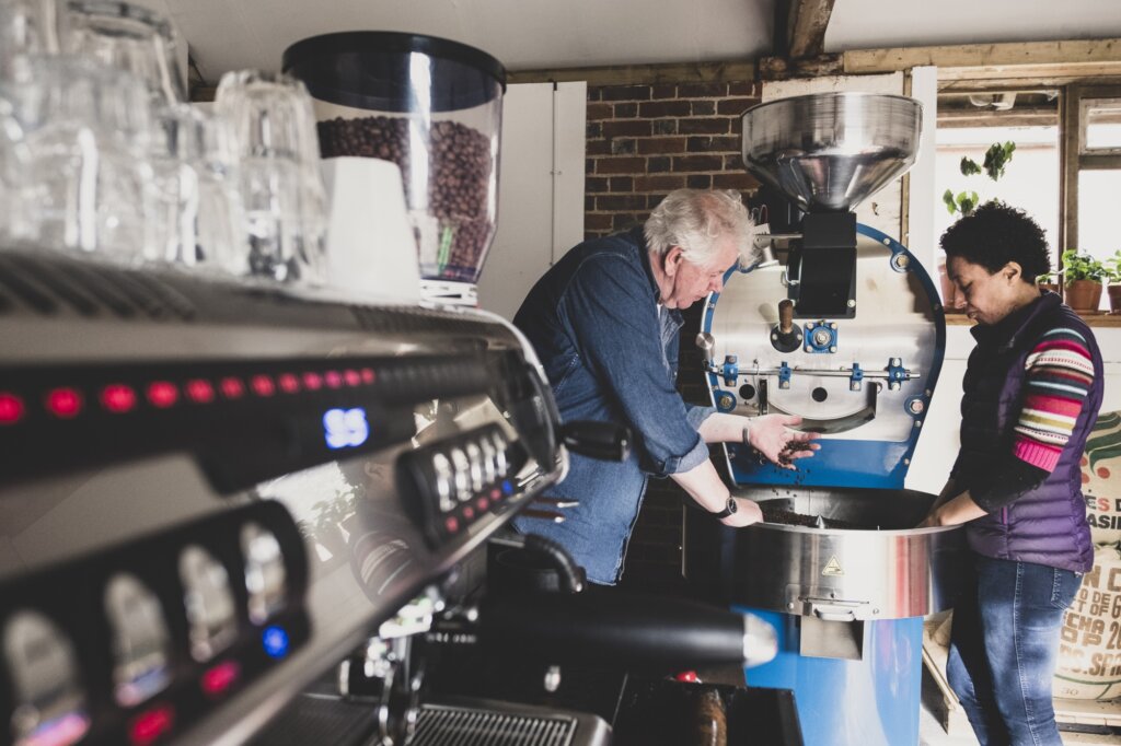 Man and woman standing next to coffee roaster, checking coffee beans.